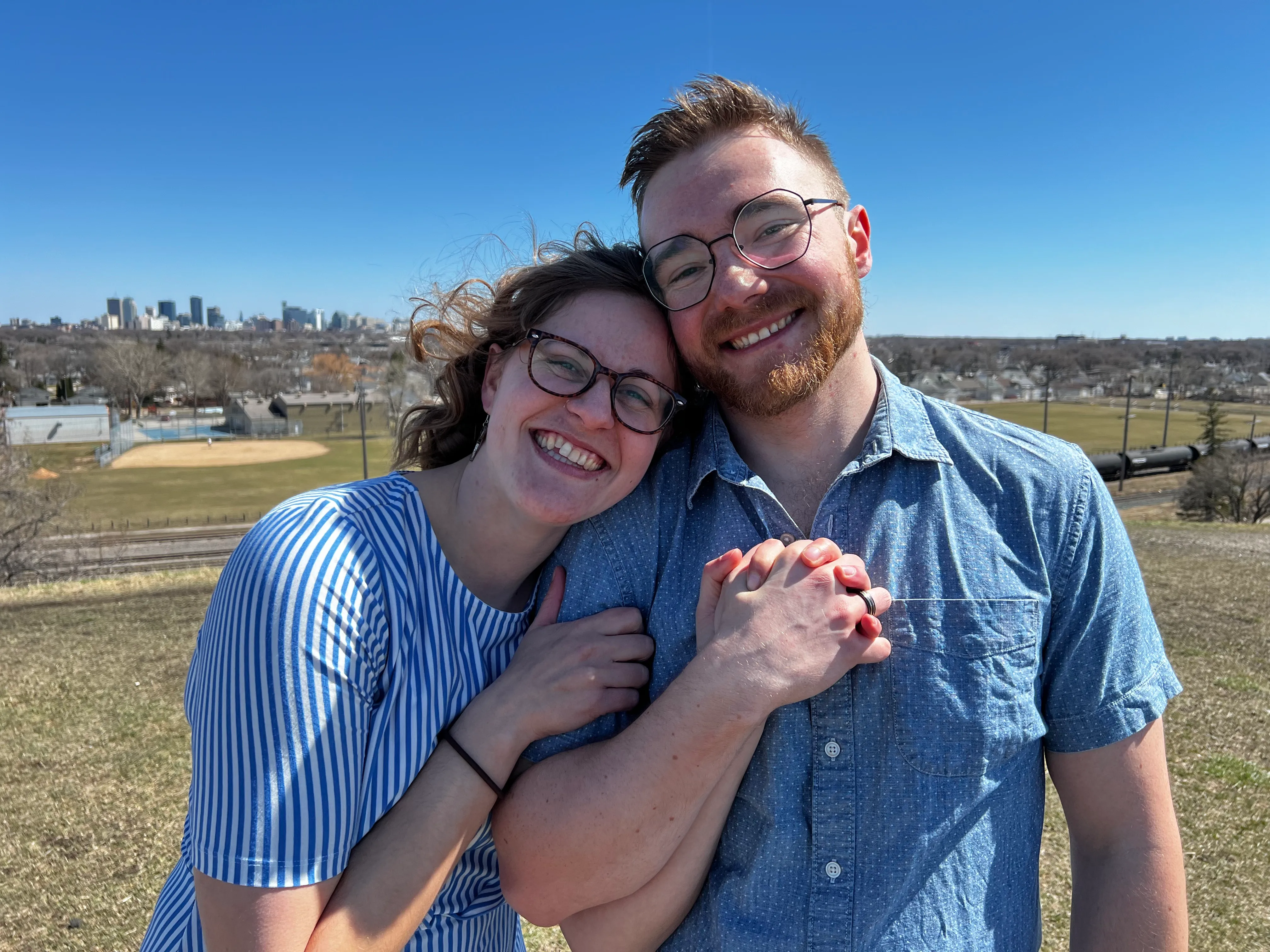 Erika and Josh stand side-by-side in on top of Westview Park's hill on the day of their engagement. The are holding hands with rings that they have just put on one another.